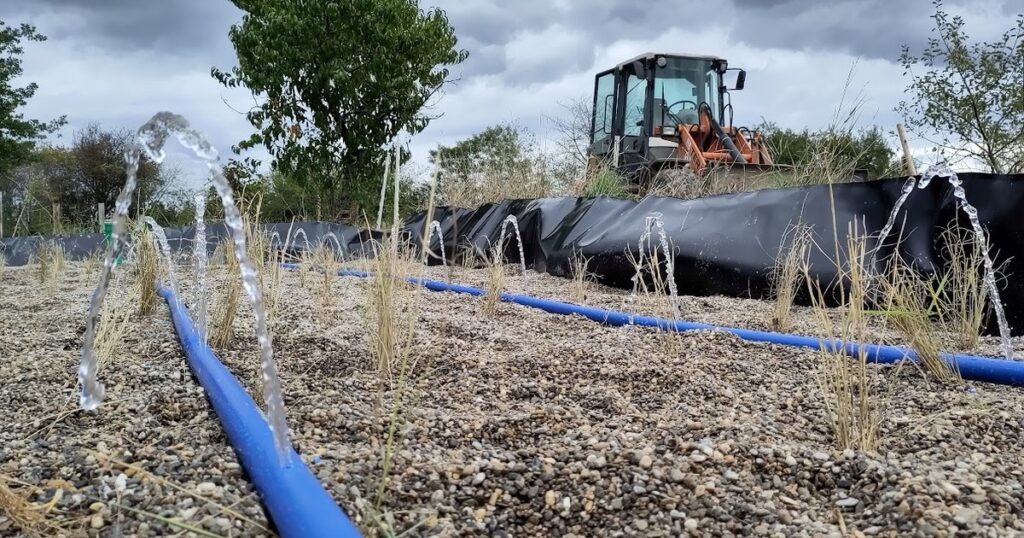 Probelauf einer Pflanzenkläranlage im Bau: Aus blauen Verteilrohren im Kies spritzen Wasserfontänen, im Hintergrund Teichfolie und ein Radlader